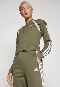 Ελαιόχρωμο αθλητικό σετ με λευκές ρίγες, ζακέτα με φερμουάρ και παντελόνι με κορδόνι στη μέση που διαθέτει λογότυπο και στα δύο. Μαλακό, ελαστικό ύφασμα.