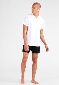 White V-neck T-shirt in soft fabric, paired with black shorts, showing a relaxed fit and short sleeves, worn by a standing model.