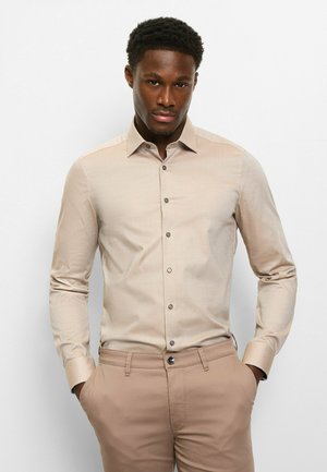 Hellbeiges Langarmhemd mit spitzem Kragen, subtiler Textur und dunklen Knöpfen, kombiniert mit beigen Hosen.