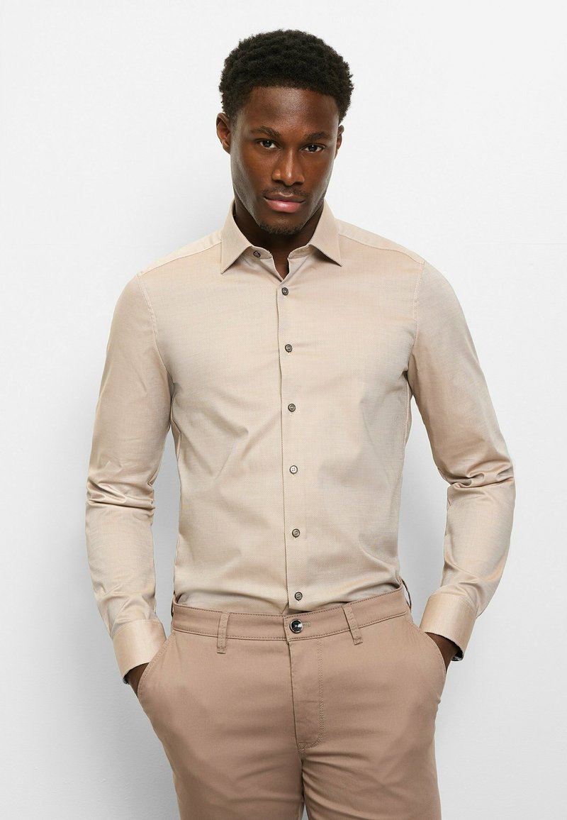 Hellbeiges Langarmhemd mit spitzem Kragen, subtiler Textur und dunklen Knöpfen, kombiniert mit beigen Hosen.