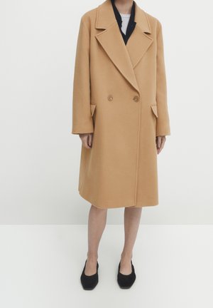 Femme portant un manteau en laine couleur chameau, croisé, avec de larges revers, des chaussures noires, et un haut blanc et noir superposé en dessous.