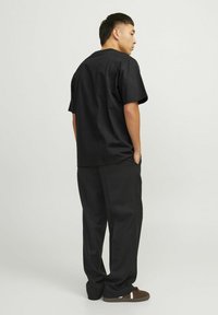 Camisa negra de manga corta combinada con pantalones negros sueltos y sneakers marrones, vista desde la parte posterior, contra un fondo neutro.