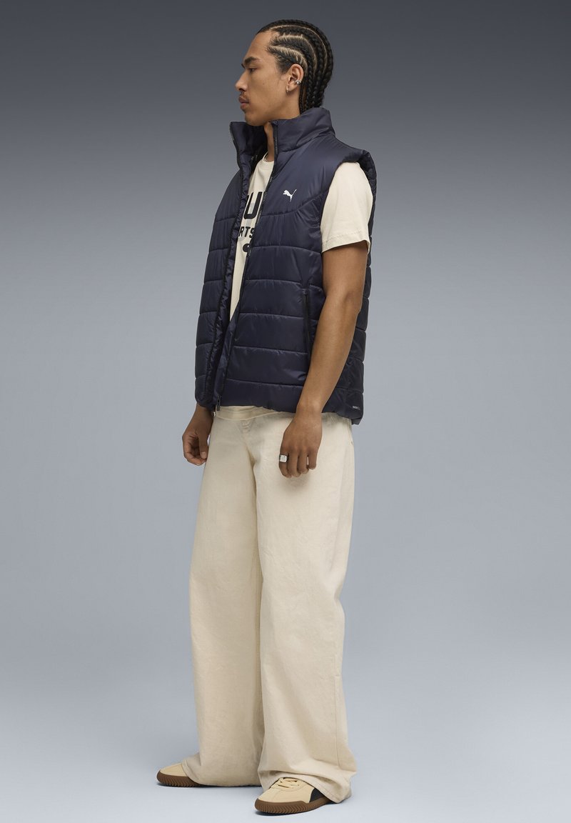 Marineblå puffer vest med lynlåslommer, båret over en lys t-shirt. Kombineret med løse kremfarvede bukser og beige sko med sorte accenter.