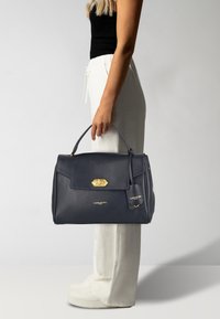 Borsa a mano in pelle blu navy con un design strutturato, hardware dorato, manico superiore e una targhetta staccabile. Presenta una texture liscia e uno stile minimalista.