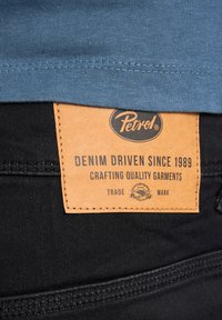 Schwarze Jeans mit einem blauen Baumwollhemdsaum, versehen mit einem hellbraunen Lederpatch, auf dem "Petrol" und "DENIM DRIVEN SINCE 1989" steht. Sichtbare Nähdetails.