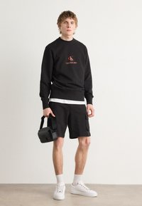 Felpa nera con logo rosso, abbinata a pantaloni corti neri con tasche laterali. T-shirt bianca sotto e sneakers bianche, mentre tiene una borsa nera.