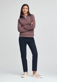 Sudadera con cremallera de color marrón claro, con puños y dobladillo acanalados, llevada sobre una camiseta de color rosa claro; combinada con pantalones oscuros y zapatillas beige.