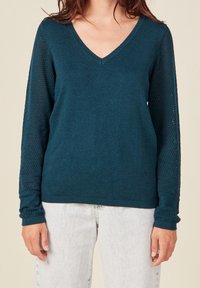 Femme portant un pull col V couleur sarcelle avec des manches en maille texturée, associé à un jean délavé clair, debout devant un fond uni.