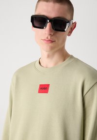 Sweatshirt vert clair avec un patch rectangulaire rouge portant le texte "HUGO". Des lunettes de soleil noires surdimensionnées complètent le look. Texture lisse, coupe décontractée.