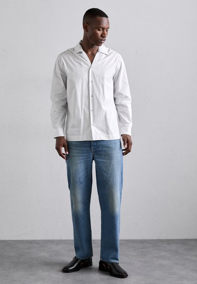 Camisa blanca de botones con cuello puntiagudo, combinada con jeans de color azul claro y zapatos negros tipo slip-on, de pie sobre una superficie gris.