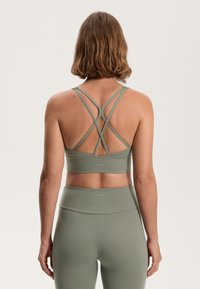 Sports bra in muted olive green, featuring crossed straps at the back and a wide band. Made from smooth, stretchy material.