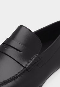 Mocassin noir en cuir à enfiler avec une lanière penny et des coutures visibles le long de l'avant-pied et des côtés sur fond blanc.