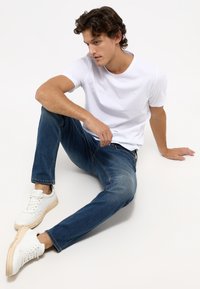 Fehér crew-neck póló, kék slim-fit farmernadrág és fehér sportcipő. A modell ül, enyhén balra néz, laza testtartásban.