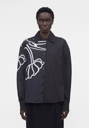 Personne portant une chemise noire boutonnée avec un grand motif floral abstrait blanc sur le côté gauche, debout devant un fond uni.