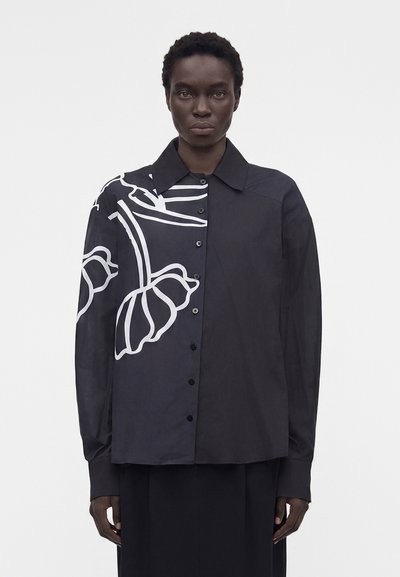 Person wearing a black button-up shirt with a large white abstract floral design on the left side, standing against a plain background.