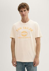Kerge virsikuvärvi puuvillane T-särk, millel on ümmargune kaelus, lühikesed varrukad ning oranž ja valge graafiline trükk koos "TOM TAILOR" logoga.