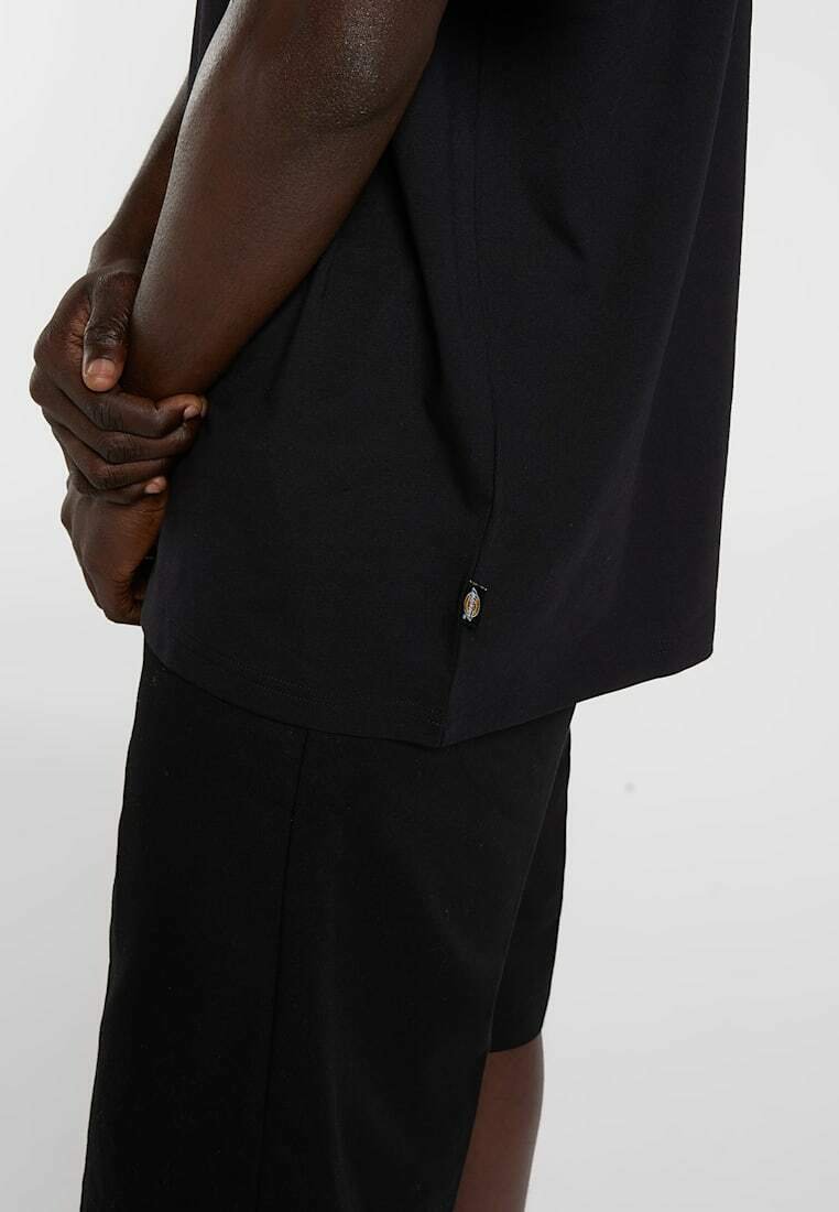 Person trägt schwarzes kurzärmeliges Hemd und schwarze Shorts, die Hände hinter dem Rücken verschränkt, steht vor einem einfarbigen hellen Hintergrund.