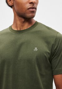 Χακί βαμβακερό μπλουζάκι με στρογγυλή λαιμόκοψη, με μικρό λευκό λογότυπο "M O P" στην αριστερή πλευρά του στήθους. Λείο ύφασμα, κοντά μανίκια.