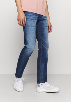 Person iført falmede blå jeans, hvide sneakers og en lyserød skjorte, stående foran en ensfarvet lys grå baggrund.