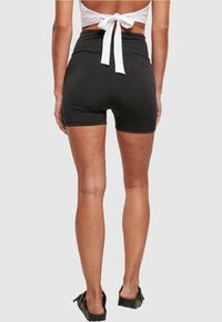 Black high-waisted shorts with a fitted design, smooth texture, and a back seam. The model wears black flat shoes with a bow accent.