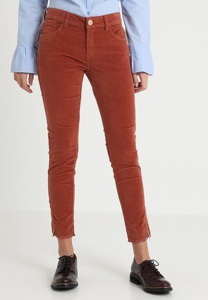 Spodnie slim-fit z sztruksu w kolorze rdzy z przednimi kieszeniami i zamkami przy kostkach, w połączeniu z niebieską bluzką i ciemnobrązowymi sznurowanymi butami.