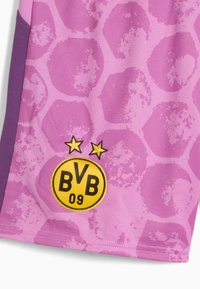 Vaaleanpunaiset urheilushortsit, joissa on teksturoitu kuusikulmainen kuvio ja purppurat yksityiskohdat. Ominaisuuksiin kuuluu pyöreä keltainen logo, jossa lukee "BVB 09" ja kaksi tähtea sen yläpuolella.
