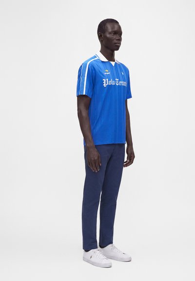Homme debout portant un polo de tennis bleu, un pantalon bleu marine et des baskets blanches sur un fond clair uni.