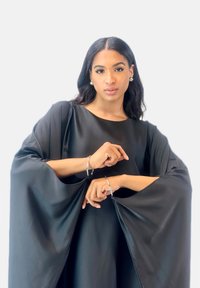 Robe noire drapée avec de larges manches, texture lisse ; mannequin portant des bracelets et des boucles d'oreilles en argent, se tenant les bras croisés.
