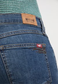 Džínové kalhoty v tmavě modré barvě s koženou etiketou s nápisem "MUSTANG True Denim." Mezi výrazné prvky patří kontrastní šití a malá červená textilní visačka.