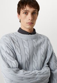 Helles blaues, kabelgestricktes Sweatshirt mit Rundhalsausschnitt, das auffällige gedrehte Muster aufweist. Darunter ist ein dunkles Hemd mit Kragen sichtbar.
