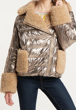 Metaliczna beżowa kurtka puchowa z dużym jasnobrązowym kołnierzem z owczej skóry, mankietami i detalami na kieszeniach, noszona na ciemnoniebieskich dżinsach.