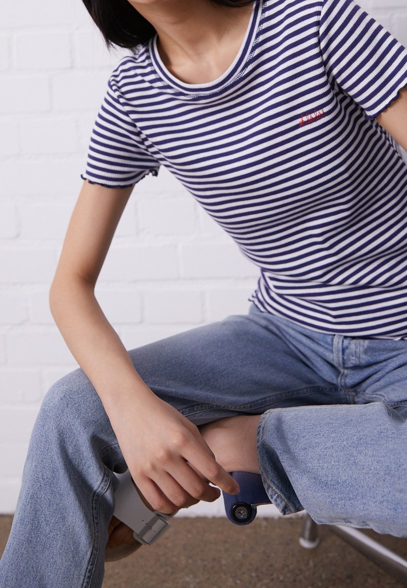 Personne portant une chemise rayée bleue et blanche et un jean bleu clair, ajustant le revers roulé du jean en étant assise.