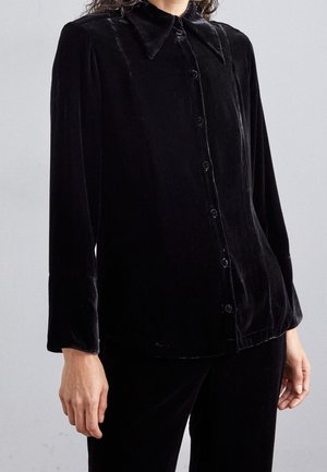 Frau trägt ein langärmliges schwarzes Samt-Hemd mit Knöpfen und spitzem Kragen, steht vor einem schlichten, hellen Hintergrund.