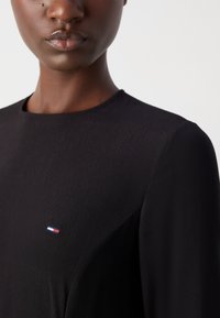 Melnā augstā kakla topa izstrādājums, kas izgatavots no struktūru auduma, ar mazu trīskrāsu logo kreisajā krūtīs. Vienkāršs, elegants dizains ar garām piedurknēm.