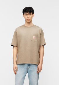 Νεαρός άνδρας με κοντά σγουρά μαλλιά, φορώντας μια μπεζ φαρδιά μπλούζα με τσέπη στο στήθος και ανοιχτόχρωμα μπλε τζιν, στέκεται μπροστά σε λευκό φόντο.