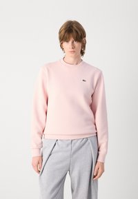Rosa sweatshirt i mjuk tyg med rundad halsringning, ribbade ärmslut och en liten grön logotyp. Tillsammans med grå, löst sittande byxor.