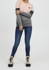 Sudadera color bloque en rosa y gris con mangas largas, tela texturizada, cuello redondo y parche de logo. Combinada con vaqueros azules y botas beig.