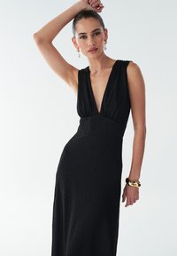 Robe noire avec un décolleté en V profond, un corsage ajusté et une jupe fluide. Tissu texturé et design sans manches. Bracelet manchette en or et boucles d'oreilles.