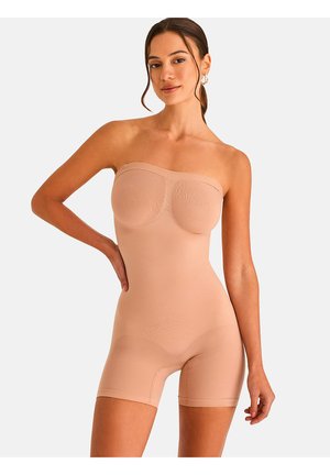 Femme portant une combinaison gainante nude sans bretelles, debout avec une main sur la hanche devant un fond clair uni.