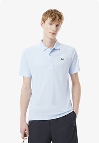 Ljusblå polo skjorta gjord av mjukt tyg, med en klassisk krage, två knappar, och en grön broderad logotyp på bröstet.