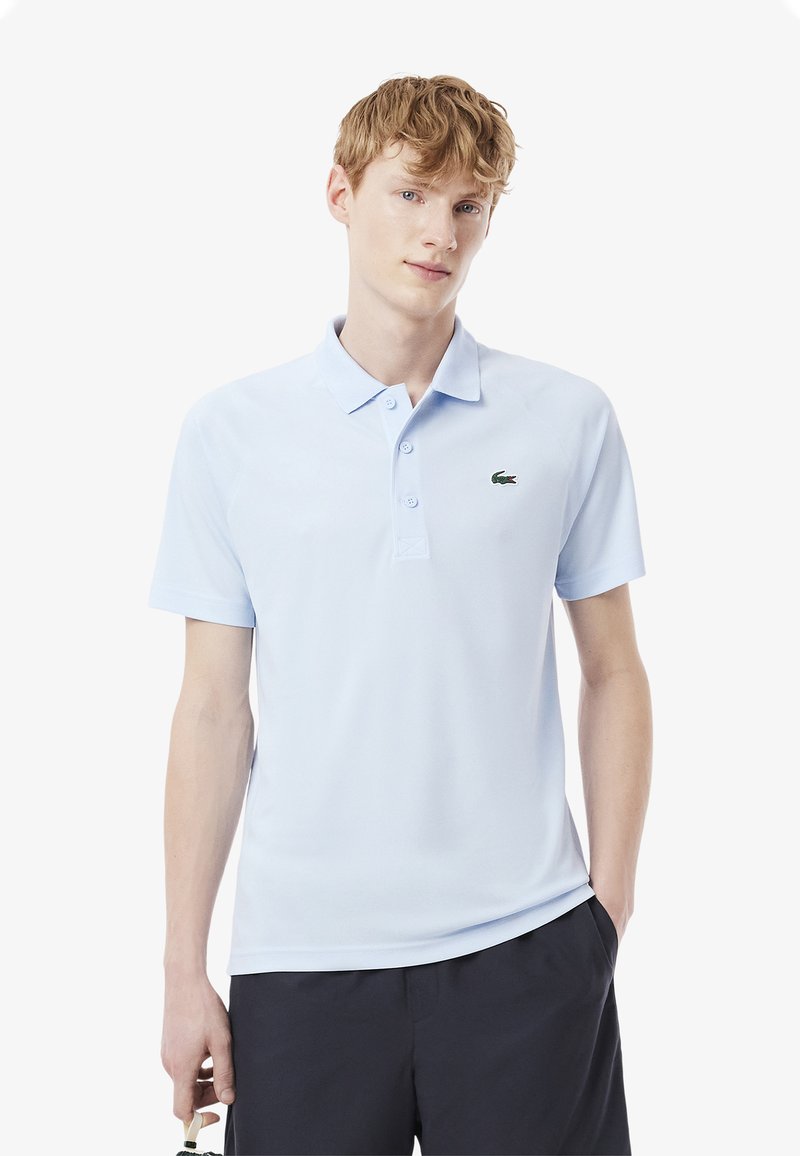 Ljusblå polo skjorta gjord av mjukt tyg, med en klassisk krage, två knappar, och en grön broderad logotyp på bröstet.