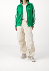 Grön lättviktsjacka med tre vita ränder längs ärmarna, kombinerad med krämfärgade baggybyxor och beige sneakers.