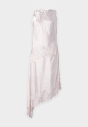 Femme portant une robe asymétrique en satin blanc sans manches avec bordure en dentelle et des sandales blanches à talons ouverts sur fond neutre.