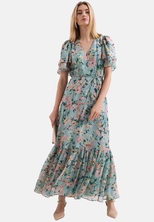 Femme portant une robe portefeuille fleurie bleu clair avec des manches bouffantes et un ourlet à volants, tenant une pochette beige et portant des talons beiges.