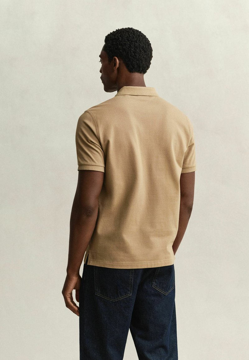 Homme aux cheveux courts et bouclés portant un polo beige à manches courtes et un jean bleu foncé, debout de dos sur un fond uni.