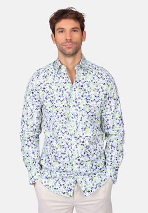 Homme portant une chemise à fleurs boutonnée avec des motifs verts et bleus, les mains dans les poches, debout contre un fond clair uni.