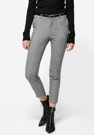 Frau trägt schwarzen Langarmtop, graue Slim-Fit-Hosen mit Hahnentrittmuster und schwarzem Gürtel sowie schwarze Reißverschluss-Stiefeletten.