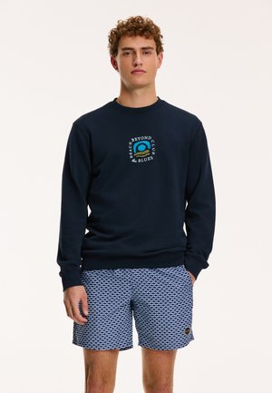 Marineblaues Sweatshirt mit einem bestickten Strandgrafik, kombiniert mit blauen gemusterten Shorts mit einem Logo-Patch. Glattes Material, Rundhalsausschnitt.