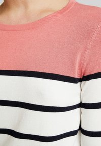 Rosa och vita randiga tröja med horisontella svarta ränder, rund halsringning och en mjuk stickad textur. Nära vy av axelområdet.