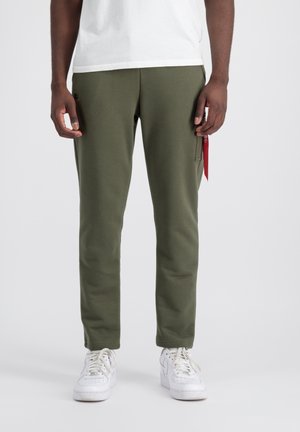 Olivgrüne Sweatpants in tapering Passform, aus weichem Material gefertigt, mit einer Seitentasche und einem roten Stoffakzent. Kombiniert mit weißen Sneakers.